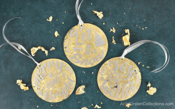 Three completed gilded gold acrylic Christmas ornaments, completed with festive decals of white vinyl and solver ribbons. Flakes of gold leaf surround the Christmas ornaments sitting on a velvet green background.