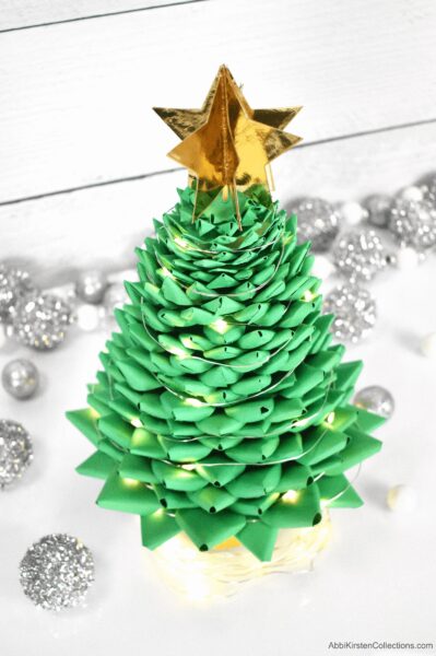 A small kelly-green paper Christmas tree, with a multisided gold star on top and fairly lights among the branches. The tree sits on a white background with silver ornaments scattered around. Create this fun and easy 3D paper Christmas tree craft for your holiday decor this year. Download the FREE SVG and PDF printable templates.