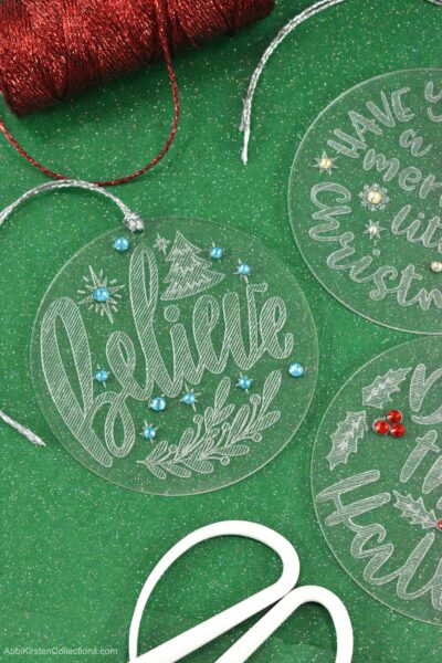 Three engraved acrylic Christmas ornaments sit on a green paper background. The ornaments are engraved with different Christmas sayings - the ornament in the center says "believe" with engraved stars, a small tree, and blue gem stones.