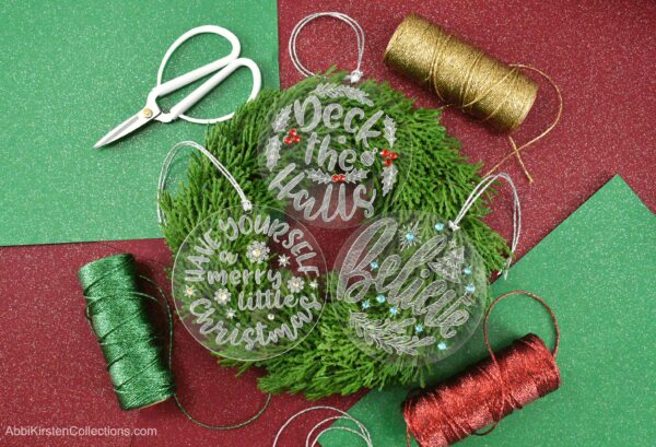 Three engraved acrylic Christmas ornaments sit on top of a balsam fir branch wreath, on a red and green paper background, surrounded by red, green, and gold spools of string. The acrylic ornaments are engraved with popular Christmas sayings - "deck the halls," "have yourself a merry little Christmas," and "believe".