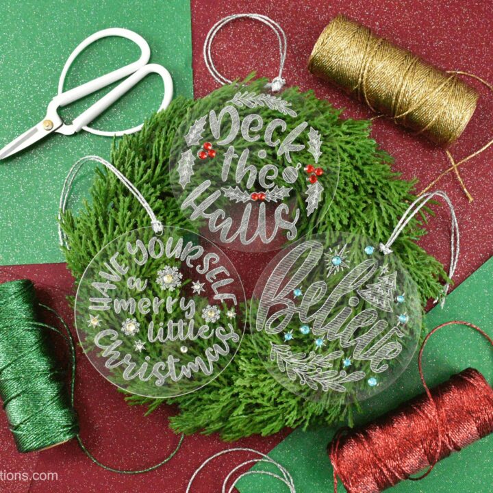 Three engraved acrylic Christmas ornaments sit on a Christmas wreath surrounded by red, green, and gold craft supplies.