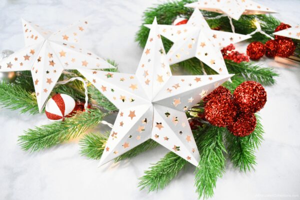 Five white paper stars create a lit Christmas garland. Pine branches and ornaments lay nearby. 