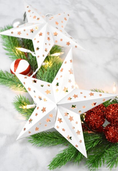 Two white paper stars with star cutouts lit by a string of white lights on a bed of festive pine branches. 