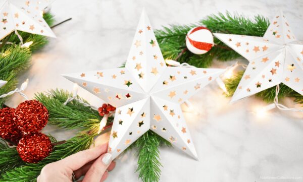 A row of white 3D paper stars creating a lit Christmas garland. Pine branches along with red and white ornaments lay nearby. 