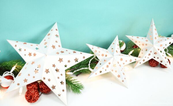 Three white paper stars in varying sizes attached by a string of white lights. The star garland is laying on festive pine branches. 
