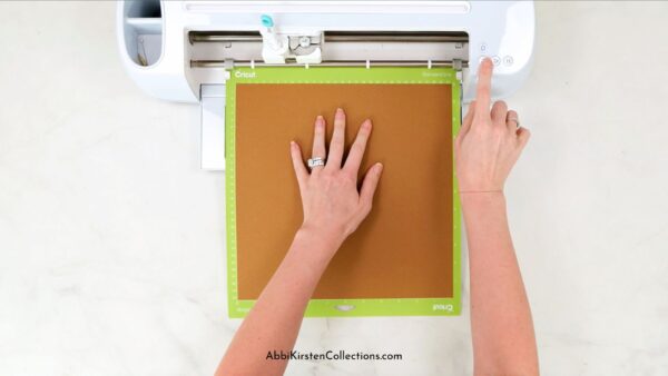 Shows Abbi's hands loading a Cricut machine with a mat that has brown cardstock on it. 