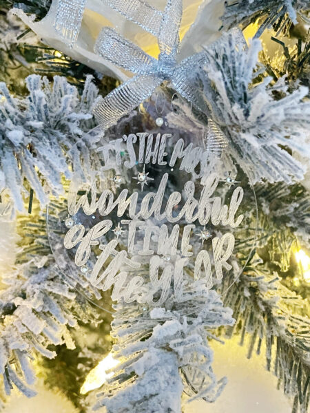 An engraved acrylic Christmas ornament hangs on a white flocked Christmas tree surrounded by silver bows and white Christmas lights. The engraving on the ornament says "It's the most wonderful time of the year" and is decorated with silver gemstones.