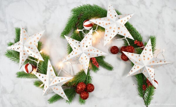 Five white paper stars lit by a string of lights. The star garland is laying on a bed of pine branches decorated with red and white ornaments. 