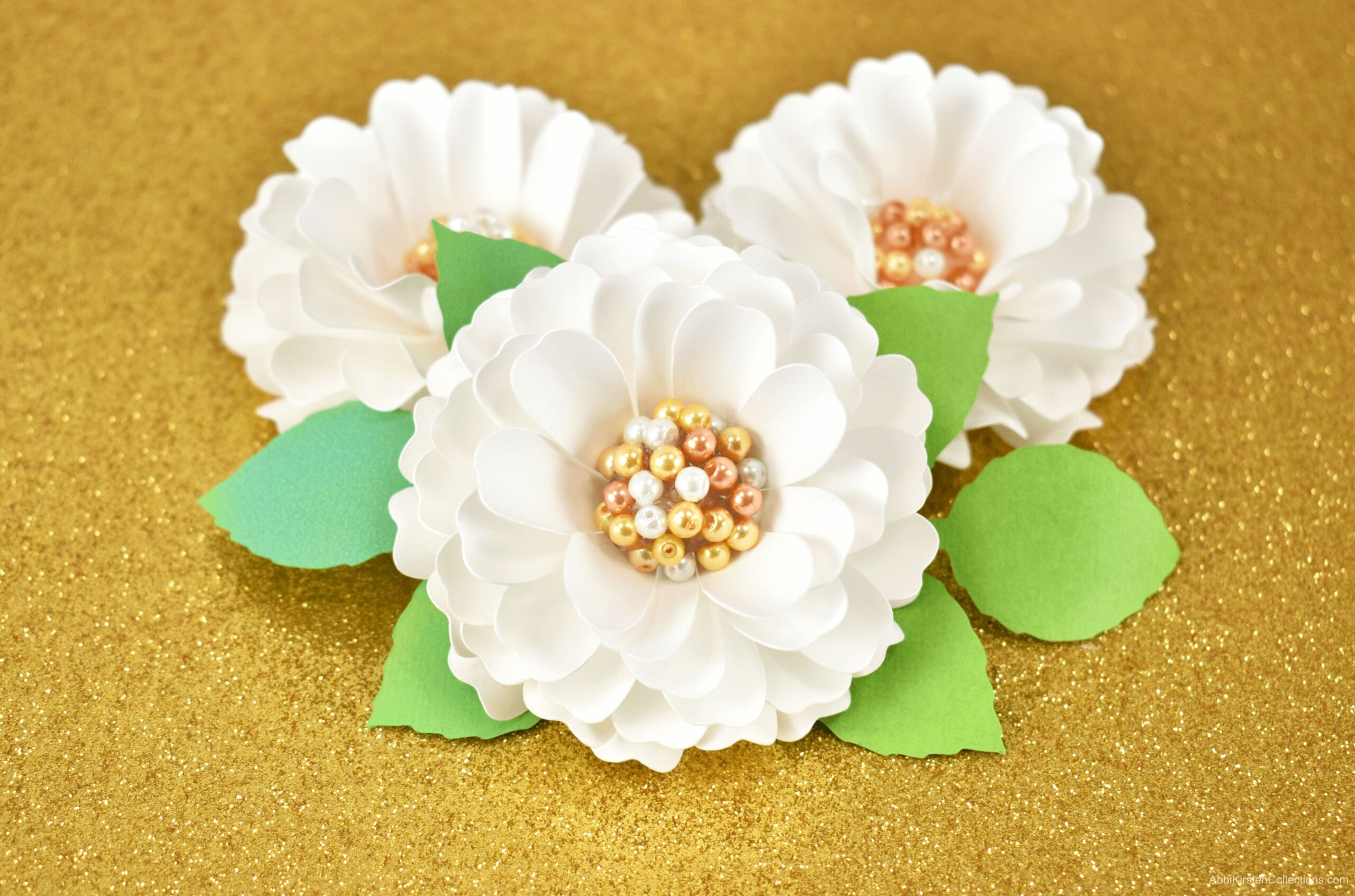 Three small white paper dahlia flowers with mini pearl centers sit on a gold background.