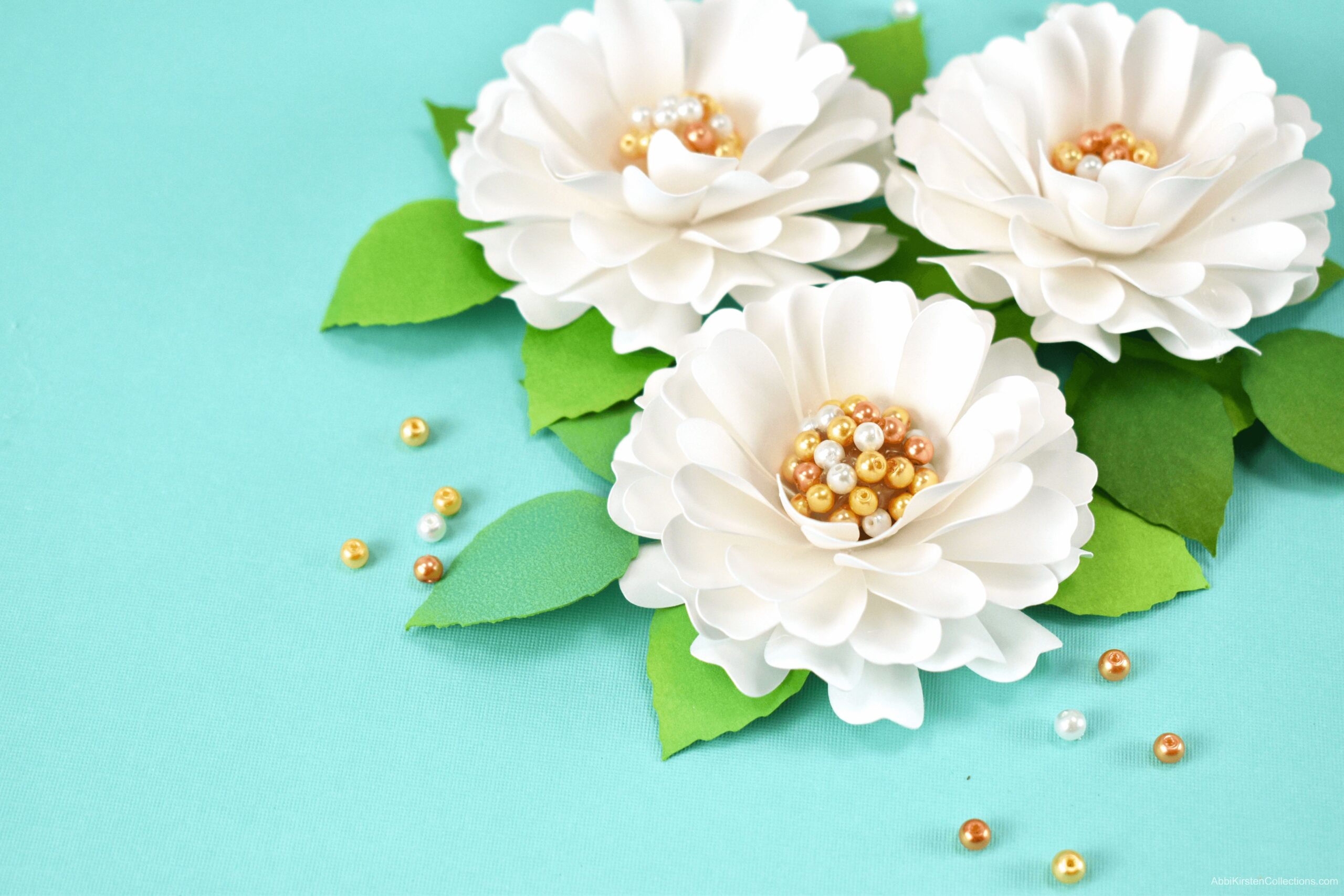 Three pearl-centered paper dahlia flowers sit on a light blue background. Gold and white pearls are scattered around the flowers.