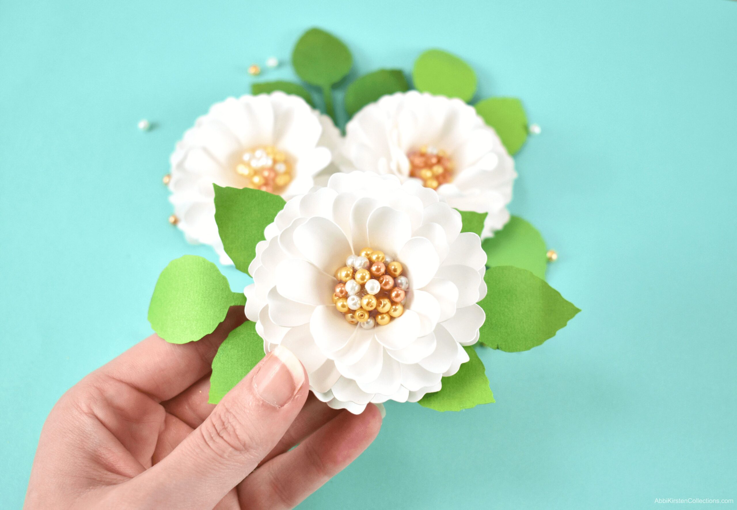 A woman holds a single small white paper dahlia flower with a pearl center. The pearls are white, gold, and brass-colored. Two more dahlias sit on a light blue backdrop in the background.