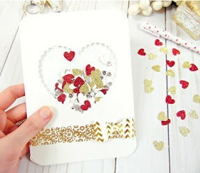 A white paper card with a heart-shaped window filled with paper heart confetti in red, gold and white. Red and gold heart confetti is sprinkled on the white wooden desk. 