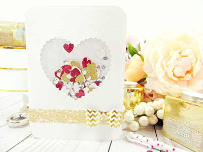 A white paper card with a gold and white bow tied near the bottom. The card has a heart-shaped window filled with gold, white and red paper heart confetti. 