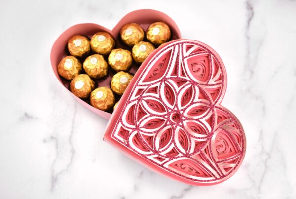 A glittery pink heart mandala paper box filled with gold-wrapped chocolates set on a marble countertop. 