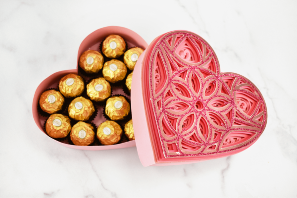 A pink paper heart mandala box filled with chocolates wrapped in gold paper. 