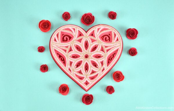 A red, pink and glitter heart mandala surrounded by red paper roses on a blue table. 