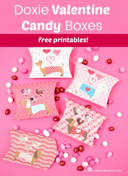 A pink graphic with "Doxie dog paper candy boxes free printables!" written in white text above an overhead view of candy boxes decorated with dogs and hearts, surrounded by candy. 