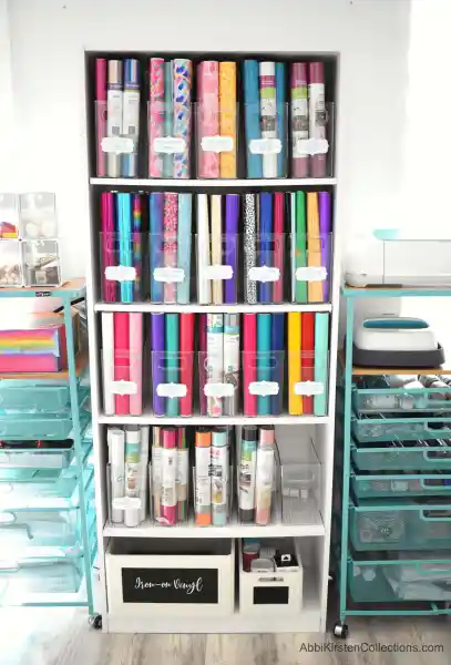 A white, tall bookshelf holds Cricut vinyl in clear storage bins organized by type. Labels make finding what you need easy and hassle-free.