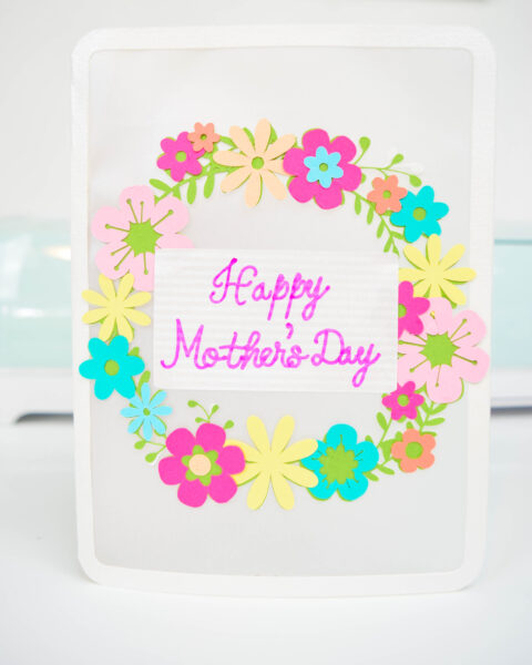 A white Happy Mother's Day card with a colorful floral wreath set on a white desk. 
