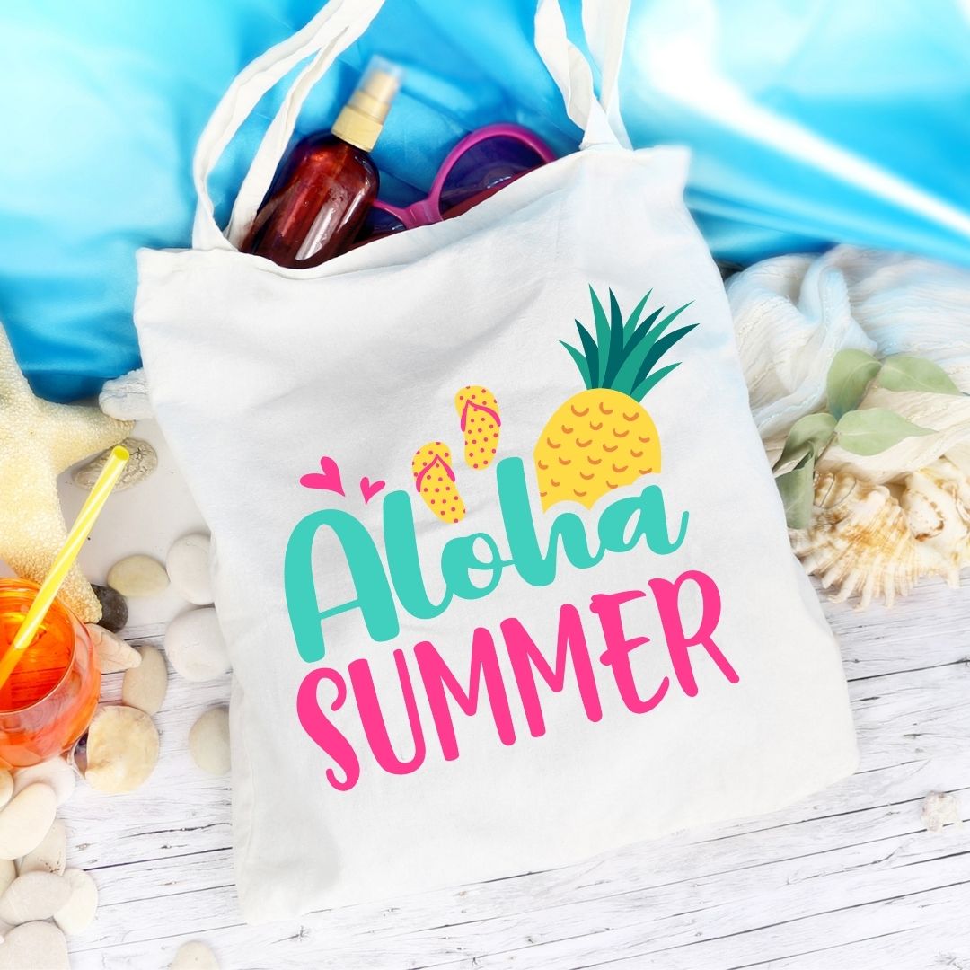 A tote bag that says "Aloha Summer" on the front with decorative pineapples lays on a wooden background surrounded by shells, sunglasses, and sunscreen.