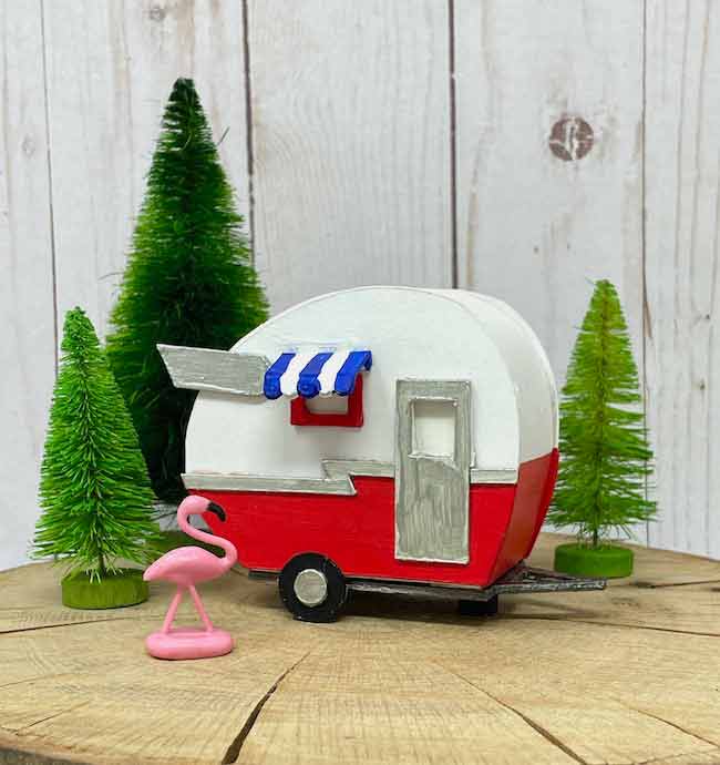 A 3D paper camper sits on a wooden table with dollhouse pine trees and a miniature flamingo lawn ornament.