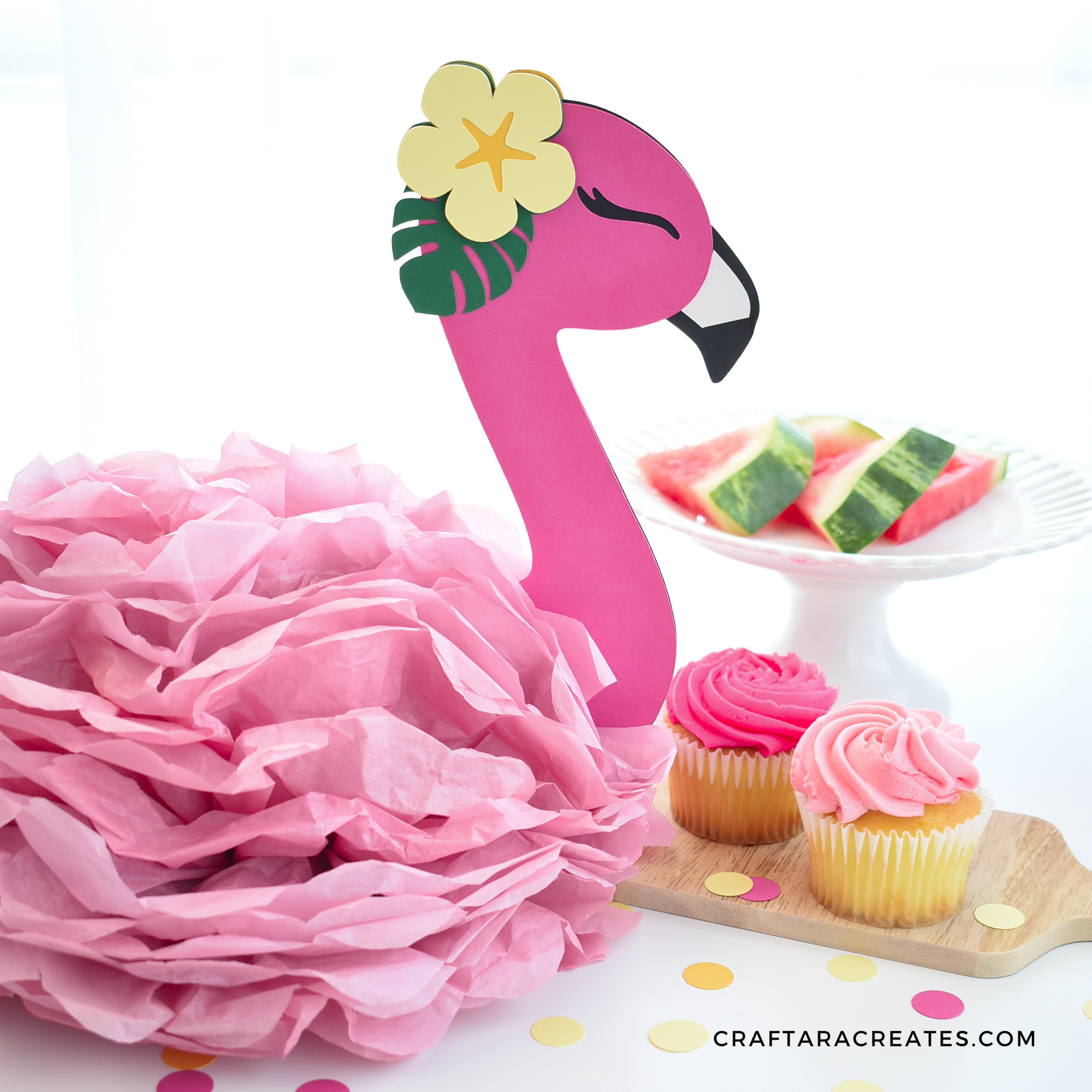 A paper flamingo with a yellow paper flower on its head sits next to a tiered plate full of watermelon slices and pink cupcakes on a white table.