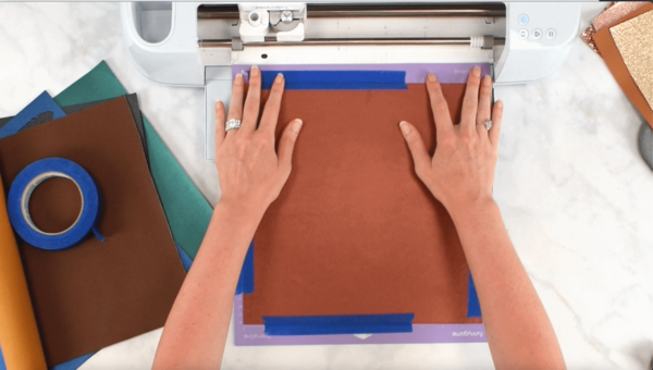 A bronze square of faux leather taped to a purple mat being inserted into a white Cricut. 