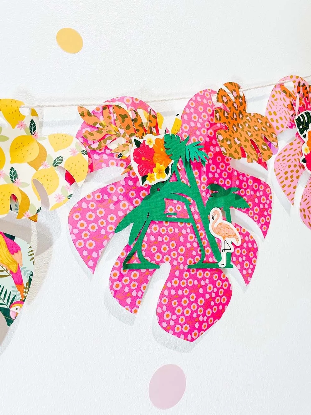 Tropical leaves with patterned paper hang as a banner on a white wall. The letter "A" is on the foremost paper leaf made of palm trees and flamingos.