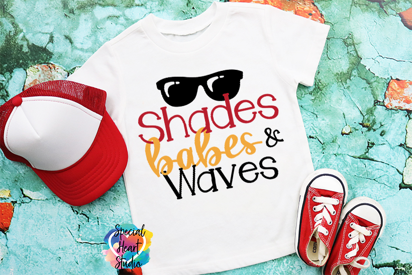 A t-shirt with a design that says "Shades, Babes, and Waves" and a sunglasses illustration lays on a green floor with red Converse shoes and a red and white hat.