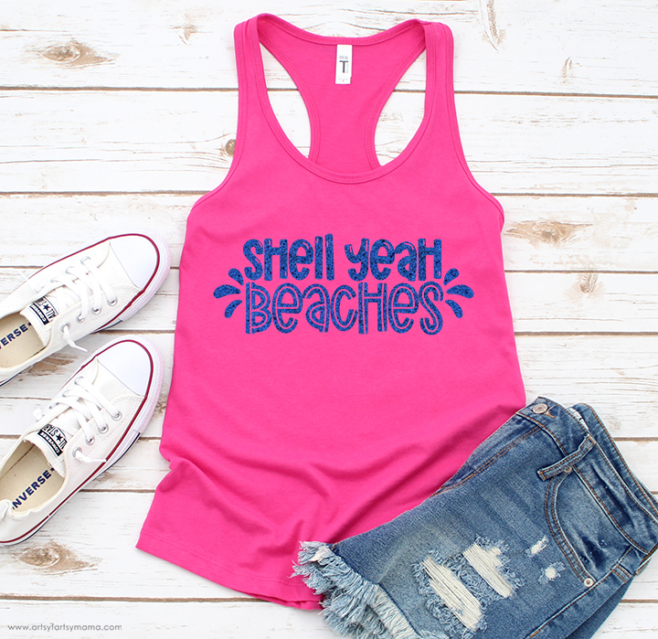 A pink racerback tank top has blue text that reads, "Shell Yeah Beaches." The top is on a white wooden background with white tennis shoes and folded denim shorts.