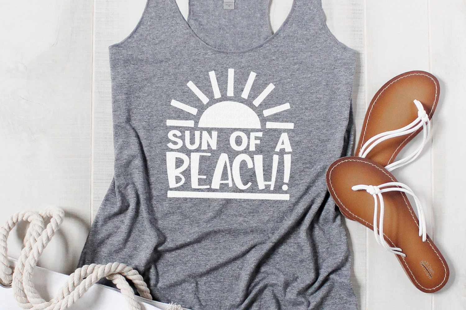 A gray tank top that says "Sun of a Beach" on the front in white vinyl. Sandals and a boat rope lay nearby.