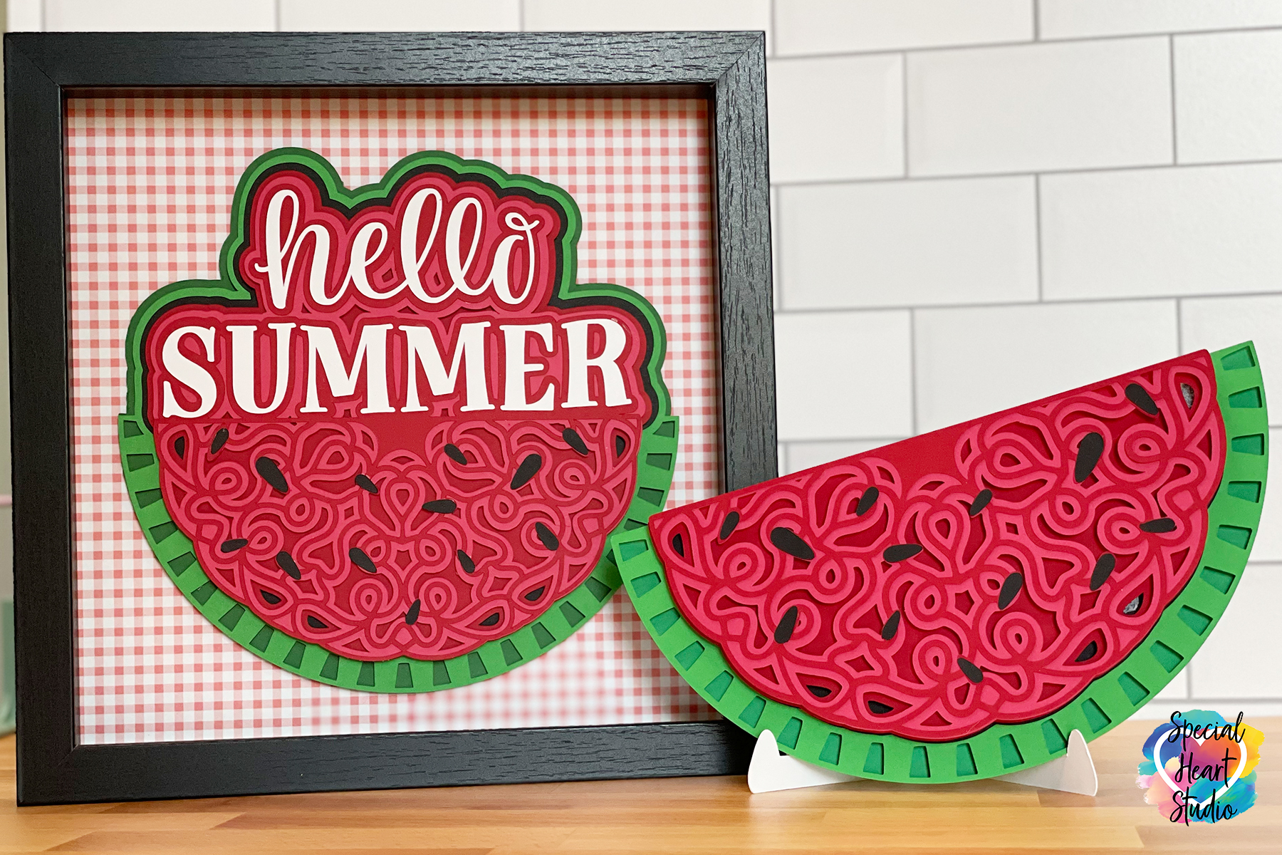 A paper watermelon made in a mandala pattern sits on a table next to a similar paper watermelon mandala in a frame with picnic checkered paper and "Hello Summer" written above.