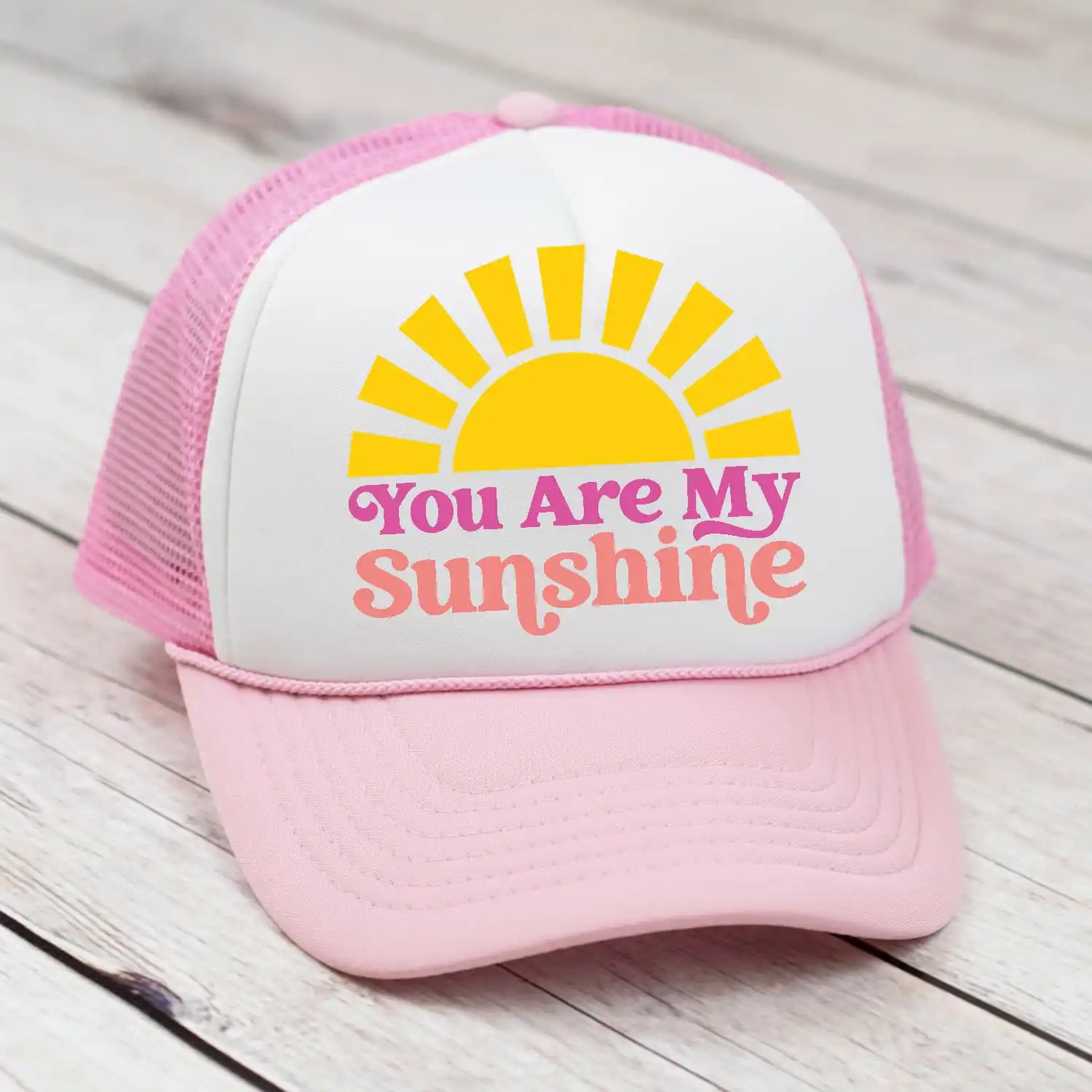 A pink and white baseball cap sits on a light grey wooden table. The words on the hat read, "You are my Sunshine" below a bright yellow illustrated sun.