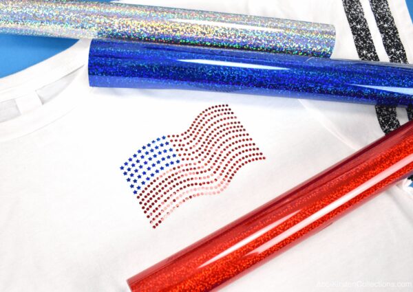 A faux rhinestone American flag made with holographic sparkle iron-on vinyl. on the pocket of a white t-shirt. The design is surrounded by rolls of Cricut holographic heat transfer vinyl. 
