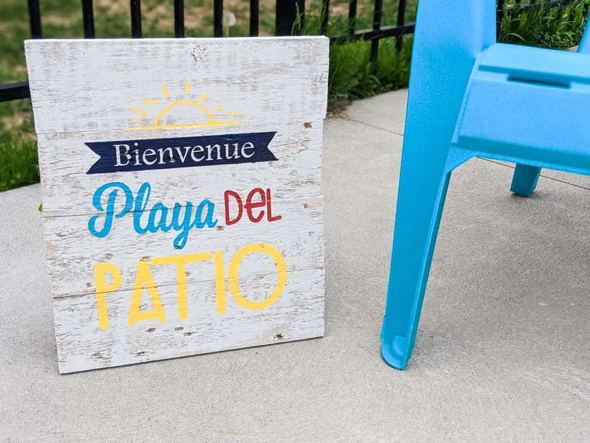 A sandwich board sign welcomes guests to the Playa Del Patio beside a blue plastic chair.
