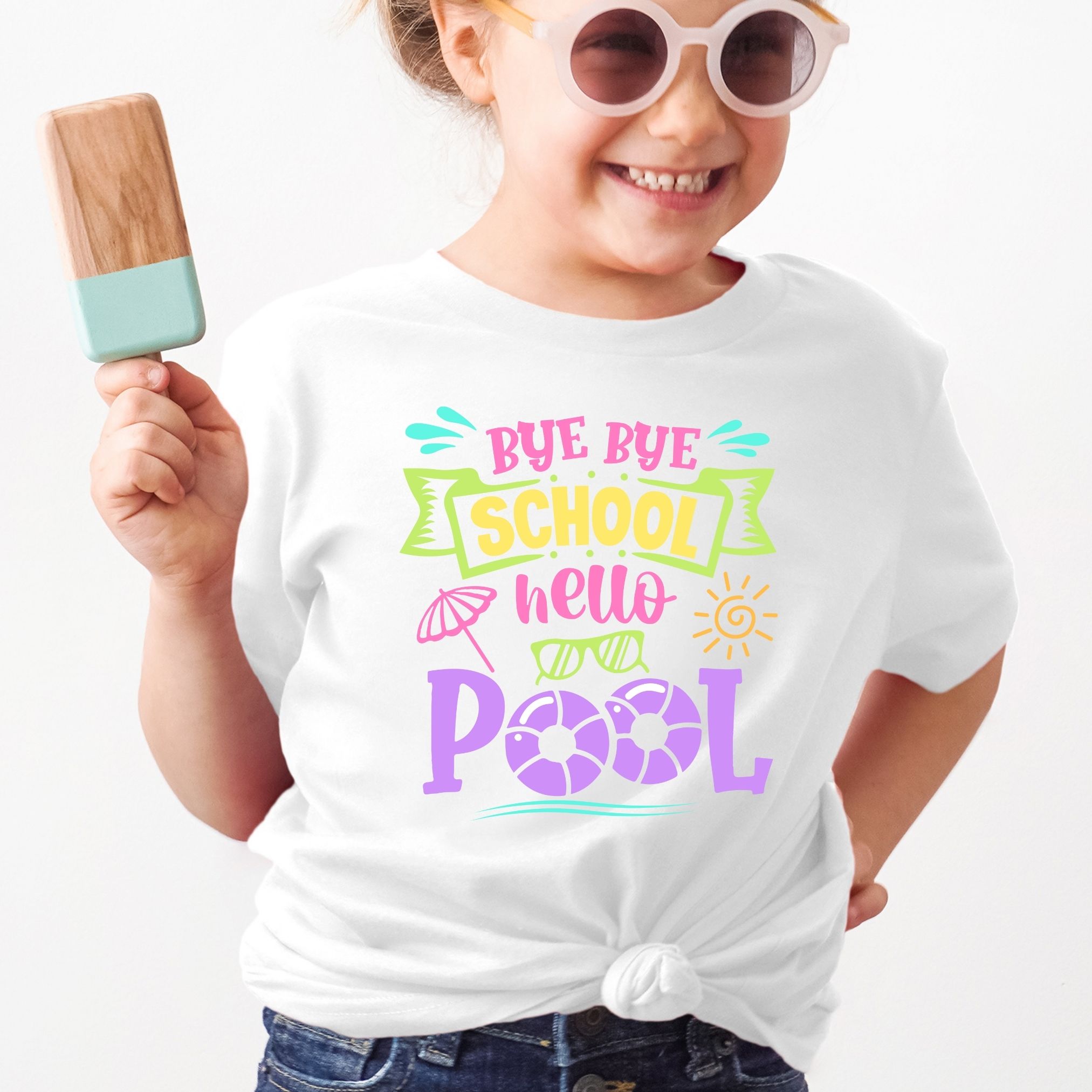 A young girl with sunglasses smiles while holding a popsicle. She is modeling a white t-shirt that says, "Bye-Bye School, Hello Pool" in bright colors.
