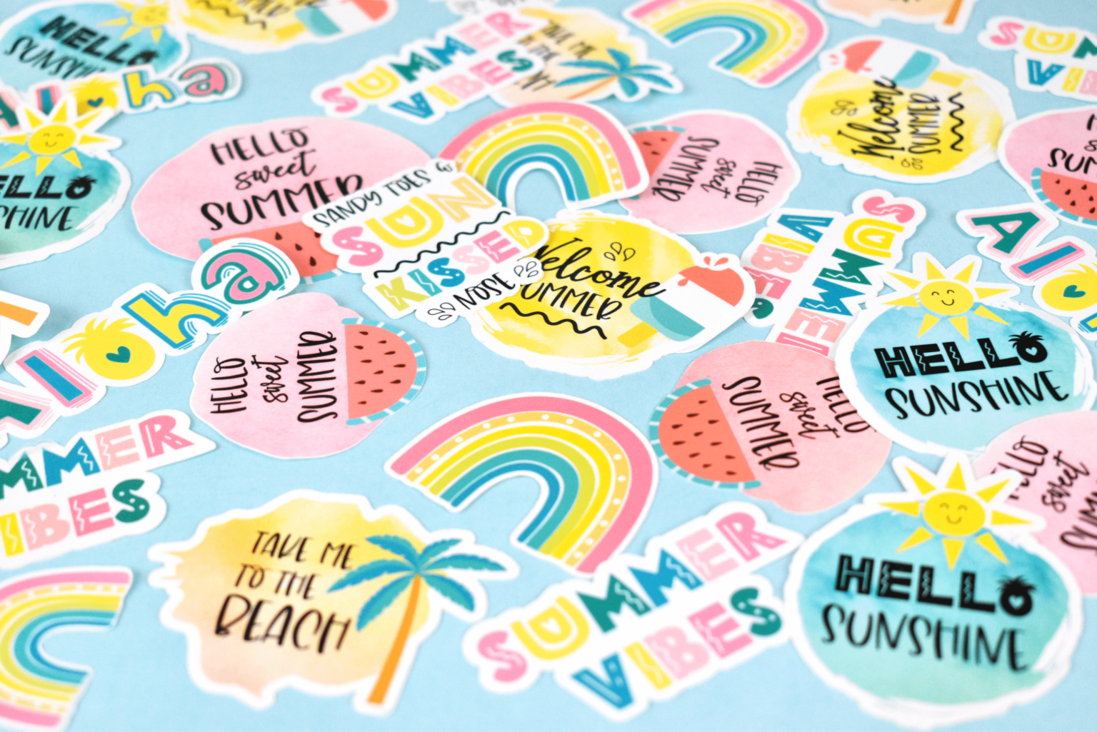 A close look at print-then-cut stickers on a light blue background. The stickers were cut using a Cricut machine. The stickers have summer-themed illustrations and quotes such as "Hello Sunshine," "Take Me to the Beach," and "Hello Sweet Summer." There are rainbows, palm trees, suns, and more on the stickers.