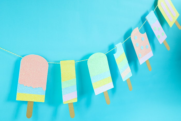 Paper pastel-colored popsicles hang on a banner against a light blue wall. This craft is available as a free summer SVG.