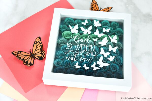 Create an easy DIY shadow box with rolled paper flowers and your Cricut. Learn the best way to apply vinyl to your shadow box and discover more cricut shadow box ideas!