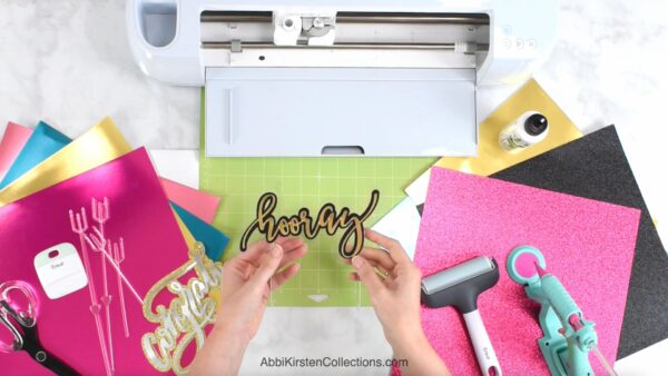 A pair of hands hold a gold and black sign that reads "hooray". The nearby workstation is filled with colorful paper, a white Cricut machine and various crafting tools. 