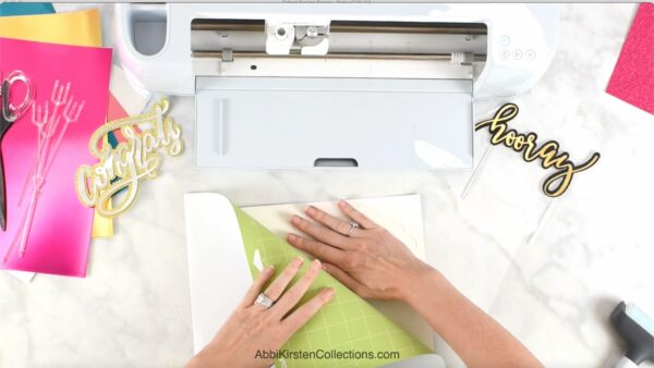 A pair of hands peeling a light green mat away from the back of a piece of paper. A white Cricut machine along with various tools lay nearby. 