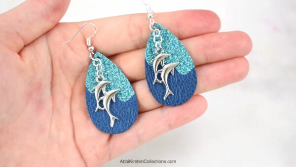 A hand holding a set of teardop-shaped turquoise and blue faux leather earrings with silver dolphin charms. 