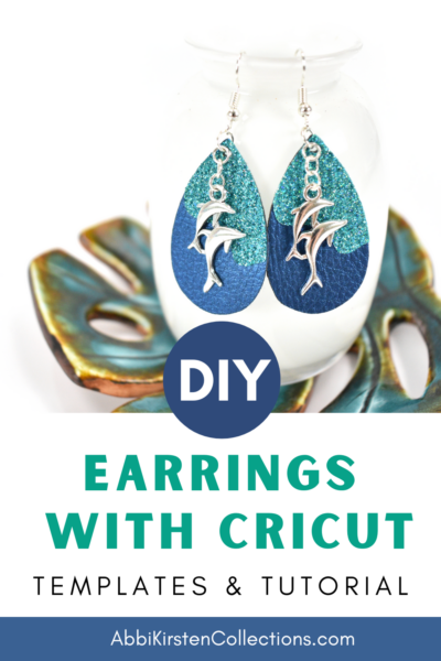 Turquoise and blue teardrop-shaped faux leather earrings with silver dolphin charms hanging from a white vase with image text overlay that reads DIY earrings with cricut templates and tutorial.