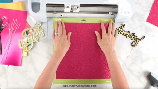 Two hands feeding hot pink paper on a light green mat through a white Cricut machine.