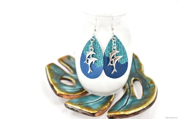Turquoise and blue faux leather earrings with silver dolphin charms hanging from a white vase set on a metallic ceramic tropical leaf dish. 
