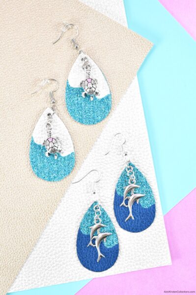 White and turquoise faux leather earrings with sea turtle charms next to turquoise and blue faux leather earrings with dolphin charms, both laying on top of sheets of faux leather. 