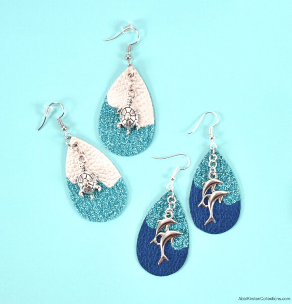 Two sets of teardrop-shaped faux leather earrings, one white and turquoise with silver sea turtle charms and the other turquoise and blue with silver dolphin charms, sitting on a light blue table. 