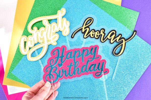 Three colorful signs that read "congrats", "hooray" and "happy birthday" laying on multicolored pieces of paper. 
