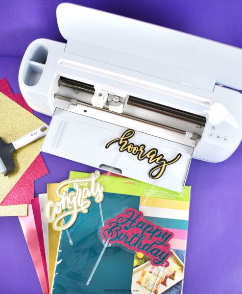 A white Cricut machine on a purple workstation. There are glitter and foil sheets of paper alongside three colorful signs that say "congrats", "hooray", and "happy birthday", respectively. 
