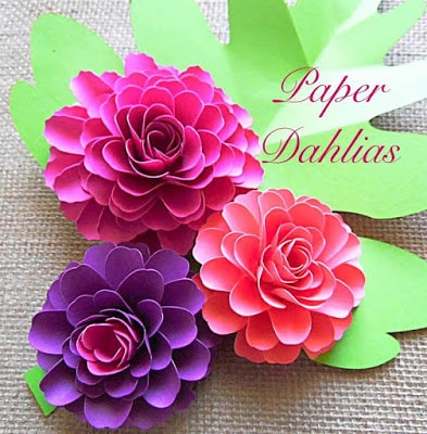 Purple and pink small paper dahlia flowers sit on bright green leaves on a burlap table covering. The words “paper dahlias” are written in the corner. 